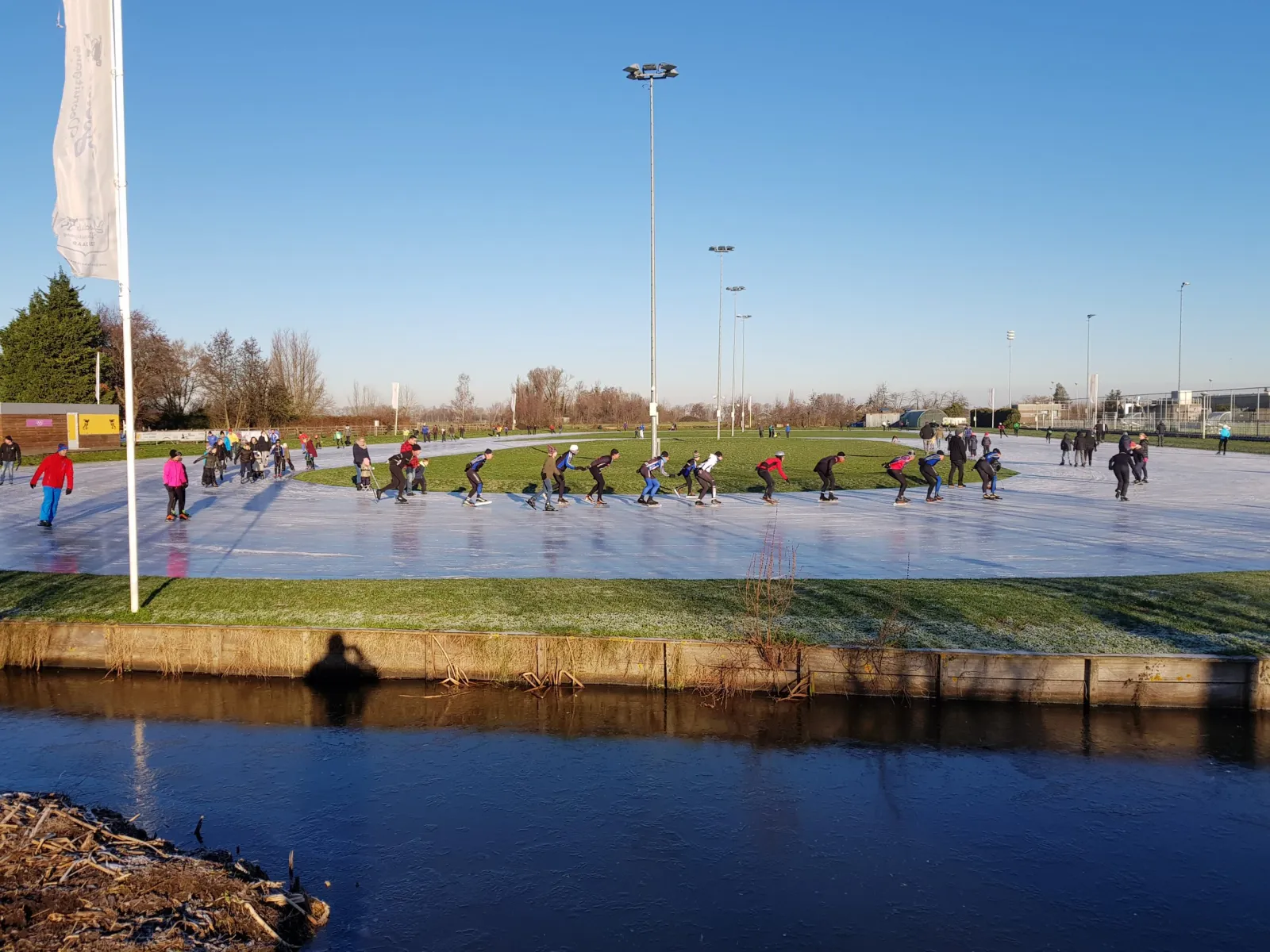 Schaatsers in een lange rij over de rechte kant van de bevroren baan van Ammerstol op een heldere dag, met lichtmasten en een vaart langs de voorrand. – IJsclub Vooruitgang Ammerstol