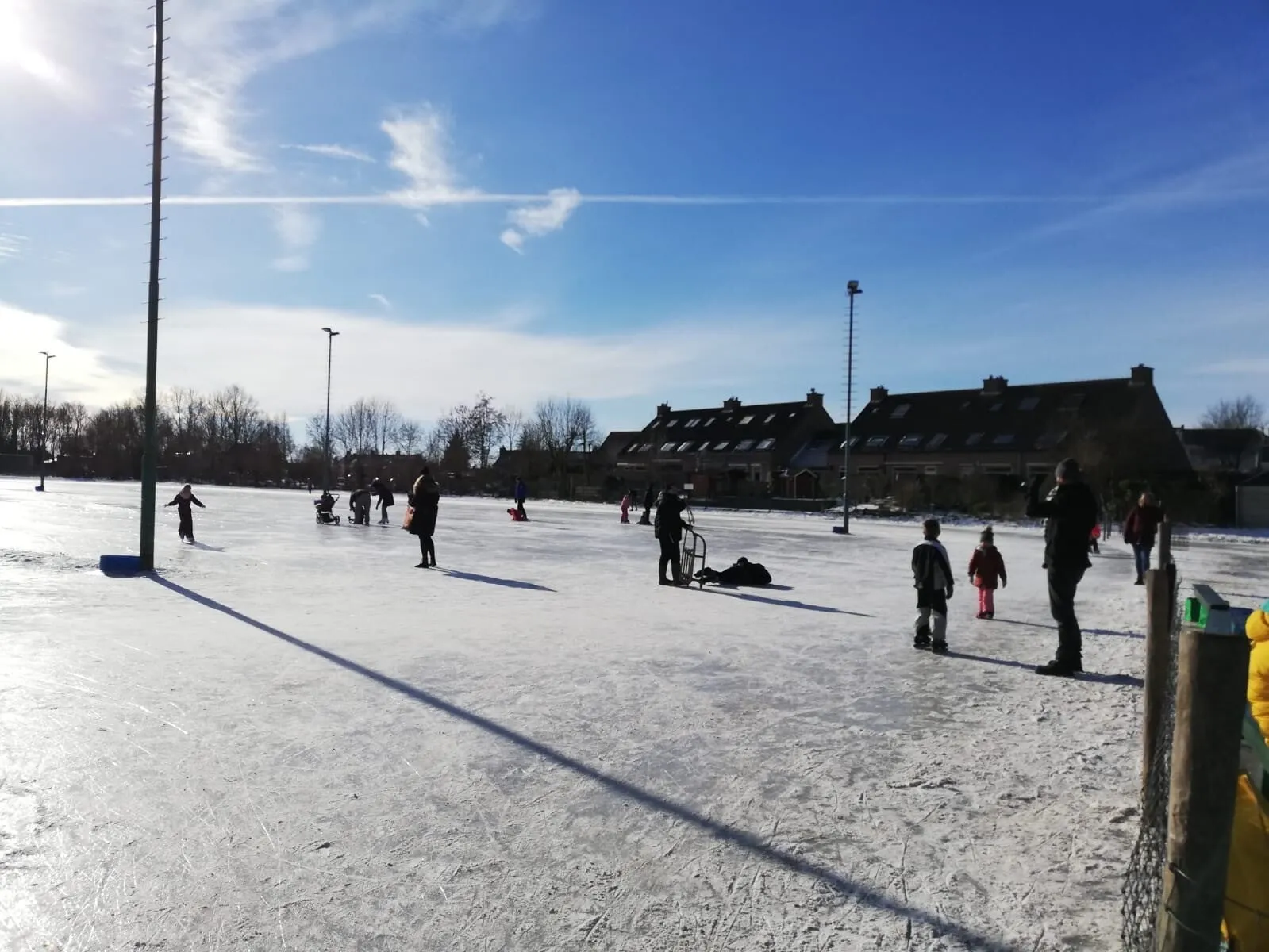 Skaters spread across the frozen Zuidland rink in winter sun, with floodlight masts and terraced houses along the far side. — IJsclub Vooruitgang