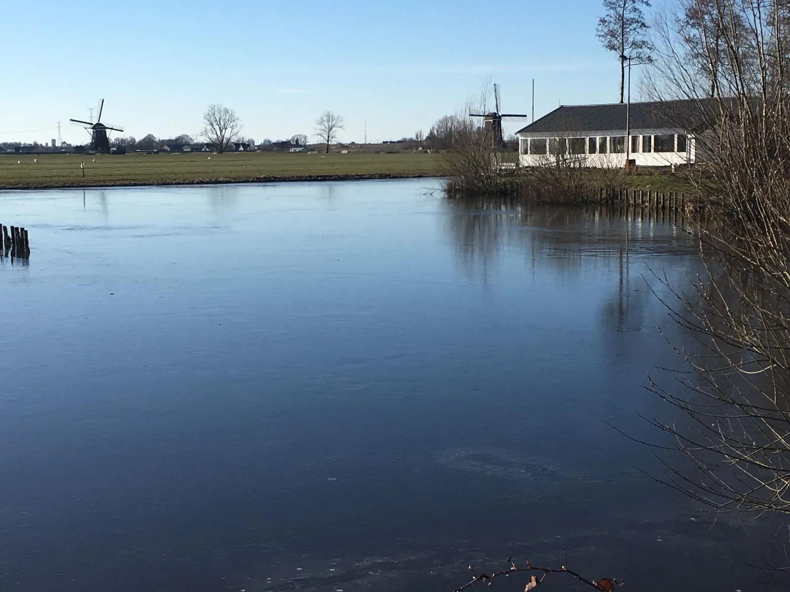 The white clubhouse of IJsclub Willem Barendts on the bank of the dark frozen rink at Streefkerk, with a windmill and open polder in the distance. — IJsbaan Streefkerk (IJsclub Willem Barendts)
