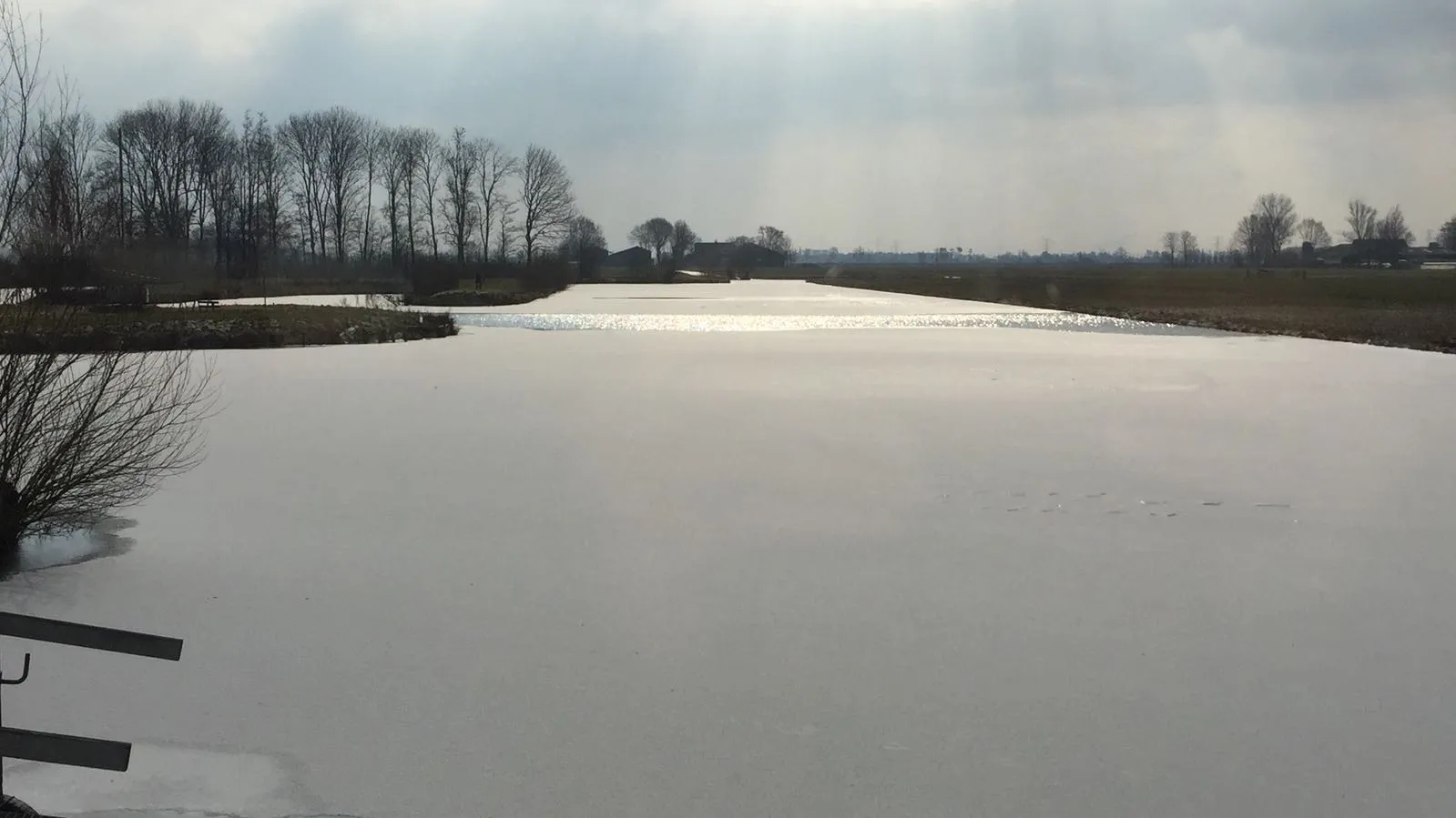 A long stretch of smooth grey natural ice through the polder at Streefkerk under a pale winter sky, with bare trees along the far bank. — IJsbaan Streefkerk (IJsclub Willem Barendts)