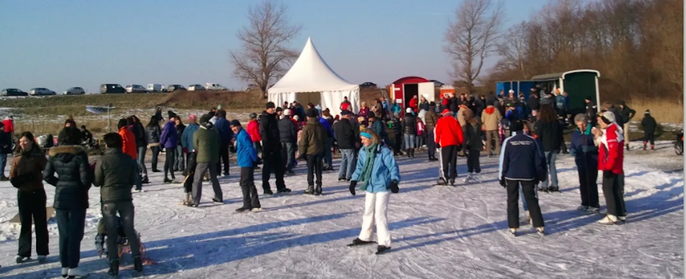 A crowd of skaters gathered on the natural ice of the IJsclub Wissenkerke at the Bokkegat on a winter day, with a white tent and sea dike behind. — IJsclub Wissenkerke (Bokkegat)