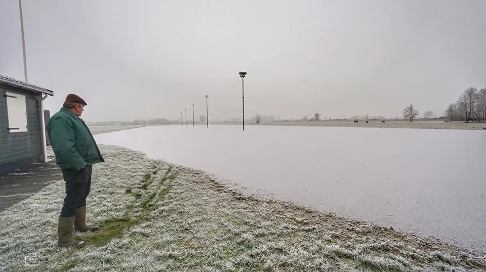 A frosty morning over the empty natural-ice rink at Zederikkade in Lexmond, with lamp posts across the ice and the clubhouse at the left edge. — IJsclub Zederik