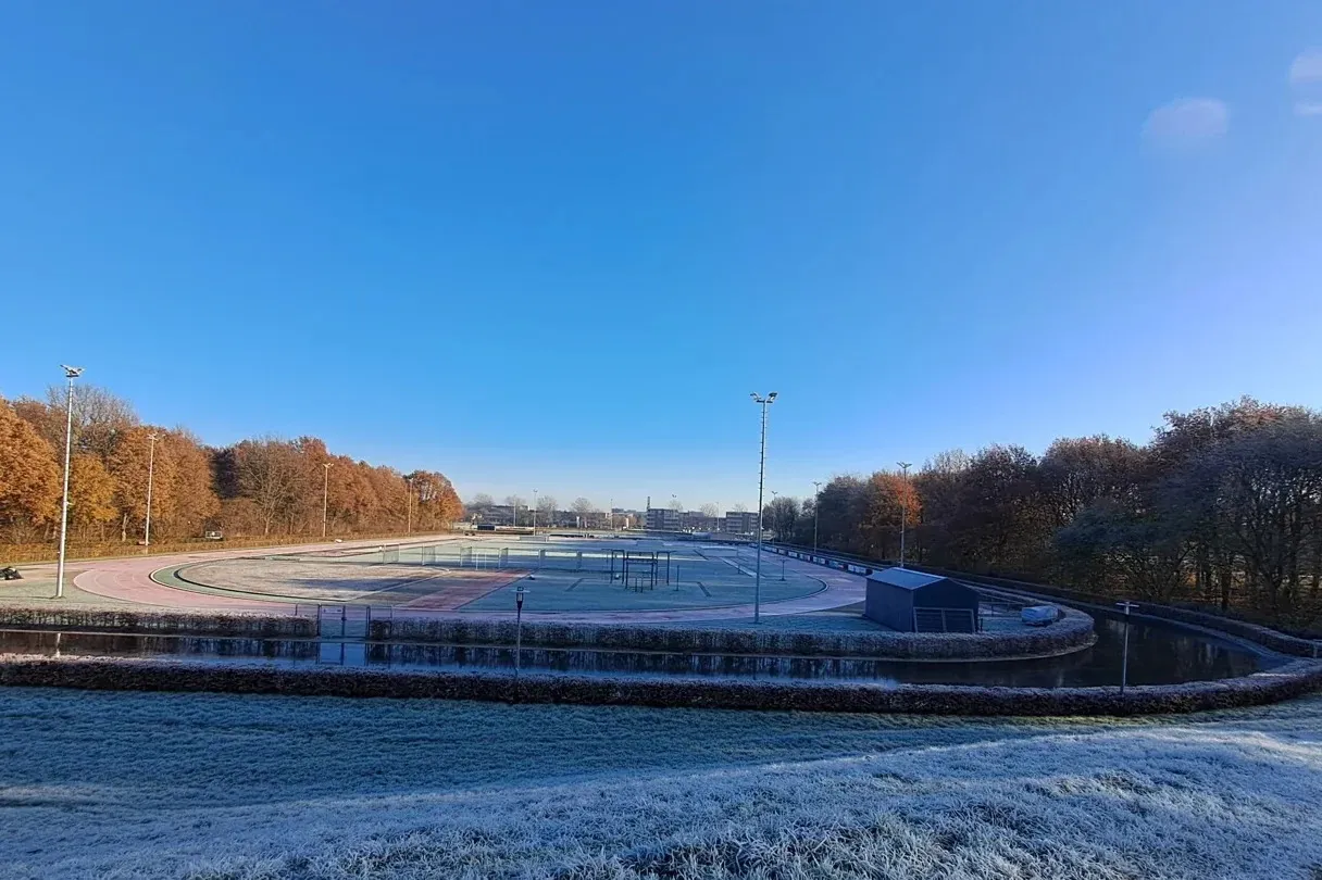 Het baanveld in Zeewolde onder een strakblauwe lucht, met lichte vorst op de grond, herfstgekleurde bomen erachter en een lichtmast aan de rand van het ijs. – IJsclub Zeewolde