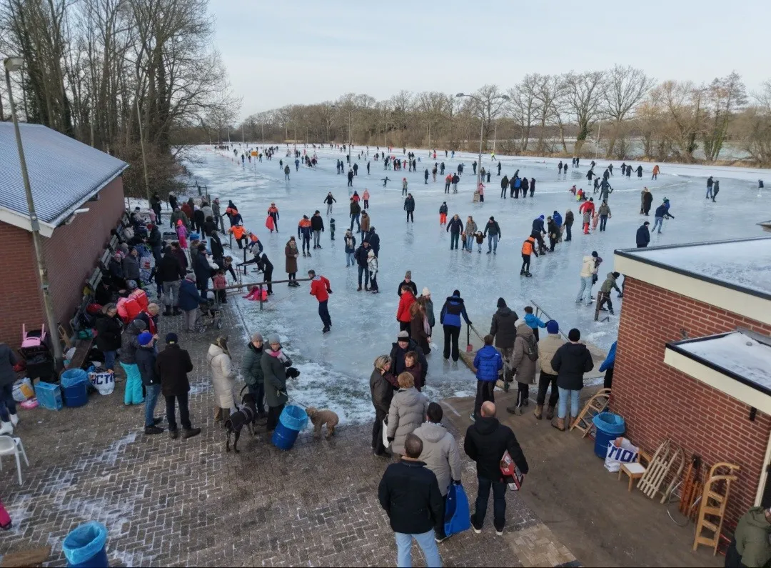 De hoek van het clubgebouw van Zenderen naast het ijs op een drukke openingsdag, met schaatsers, sleetjes en toeschouwers op het bestrate deel. – IJsclub Zenderen
