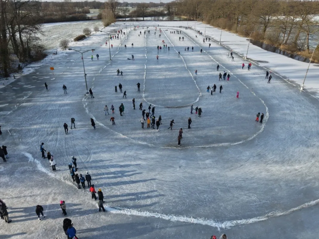 Luchtfoto van de drukke natuurijsbaan van Zenderen op 11 januari 2026, met schaatsers verspreid over de ovaal die in het ijs is afgetekend en kale bomen aan de rand. – IJsclub Zenderen