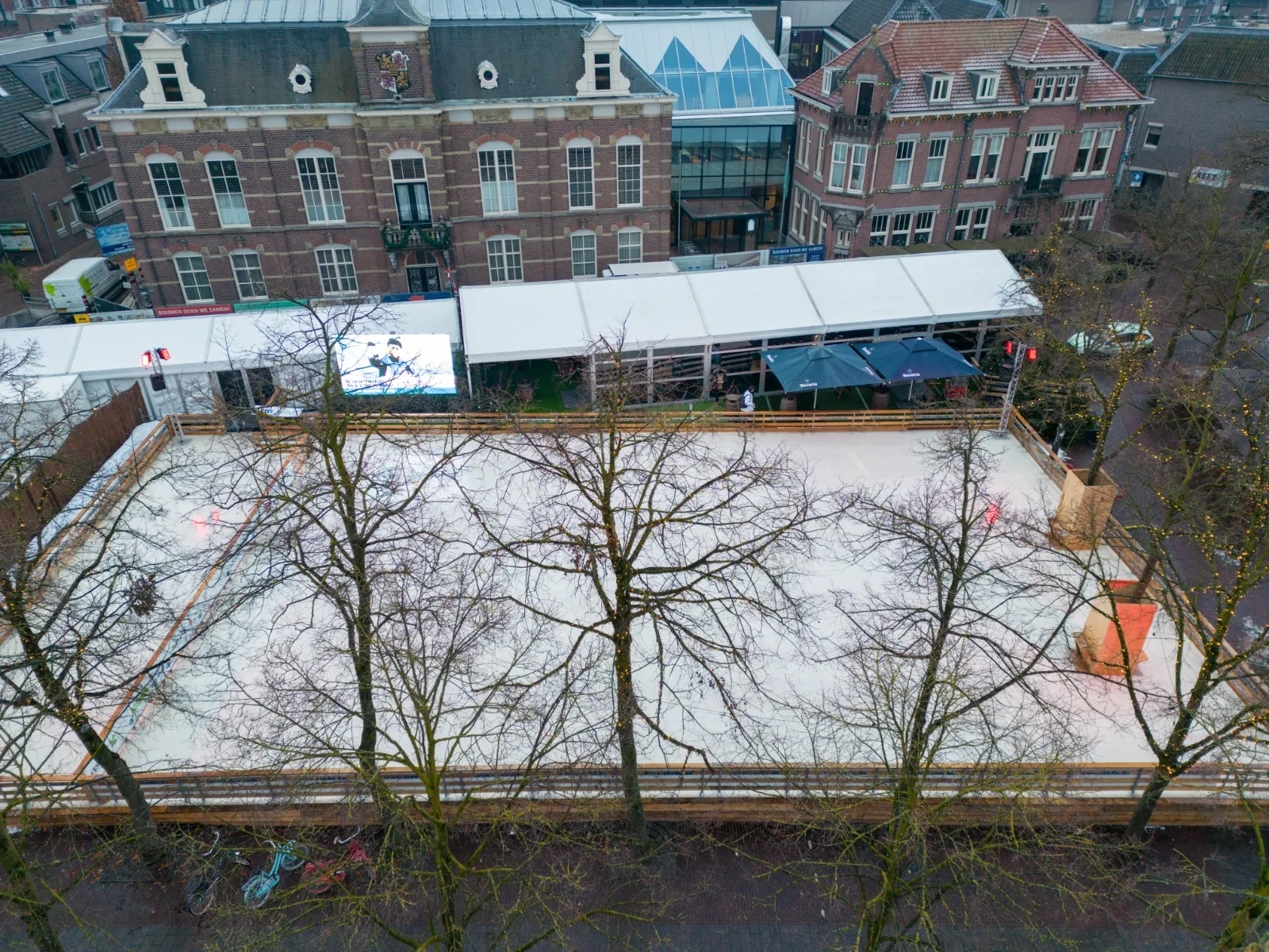An aerial view of the white temporary rink among the bare trees of the market square in Deurne, with the pavilion tents and brick buildings behind. — IJsfestijn Deurne