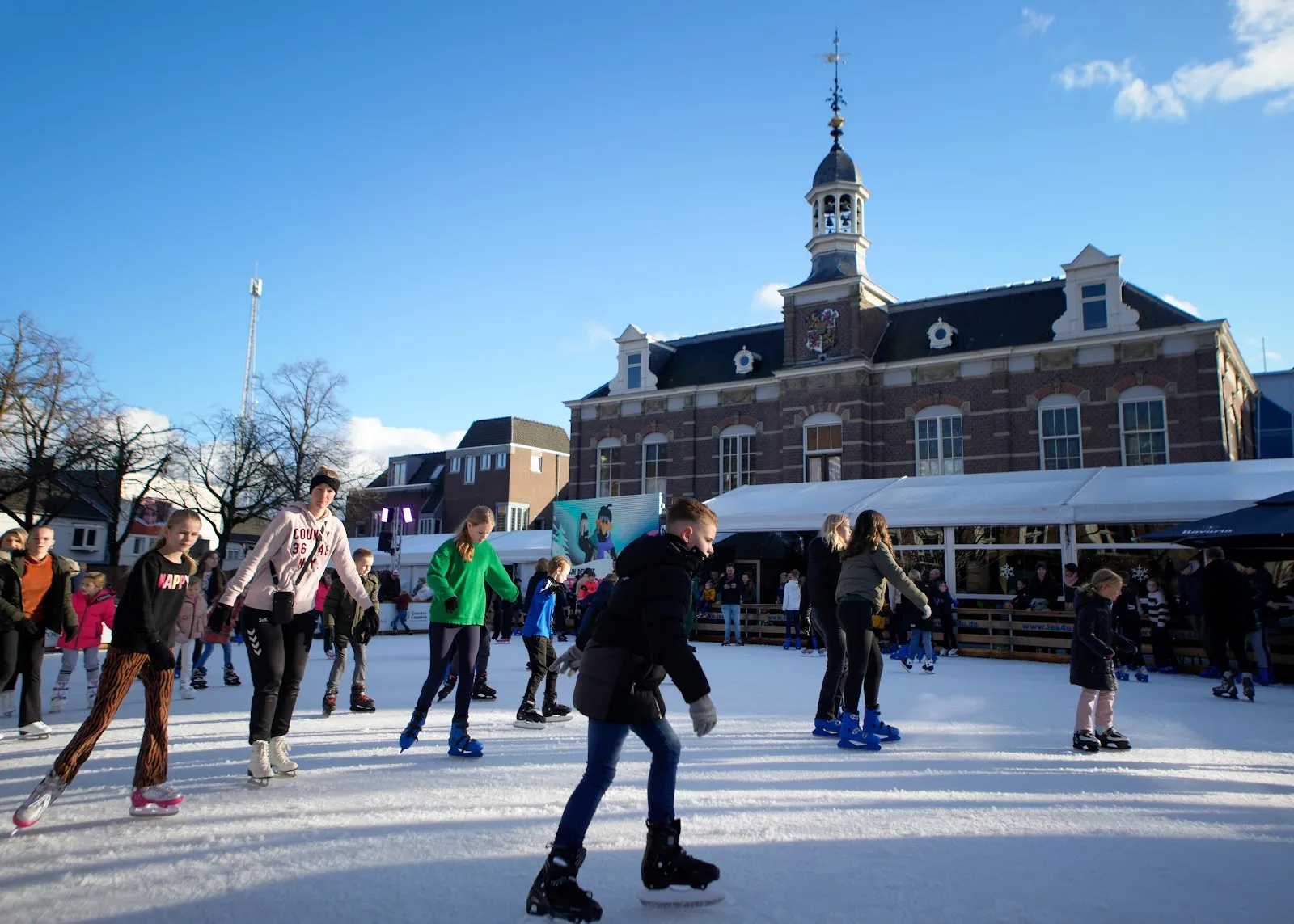 Skaters on the temporary rink in front of a historic brick building with a bell tower on a sunny winter day, with the white pavilion tent beside the ice. — IJsfestijn Deurne