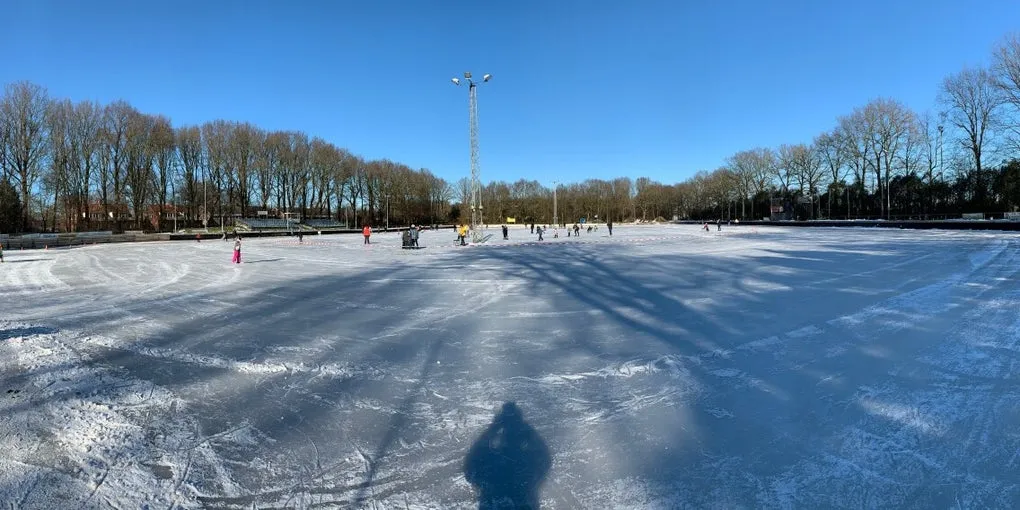 Panoramic view over the frozen De Kuip rink at Blijham on a clear winter day, with skaters in the distance and floodlight masts and trees around the edge. — IJsvereniging Blijham