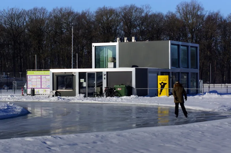 Het moderne clubgebouw naast de baan op Sportpark Zuid in Aalten op een besneeuwde winterdag, met het ijs ervoor. – IJsvereniging de Hare