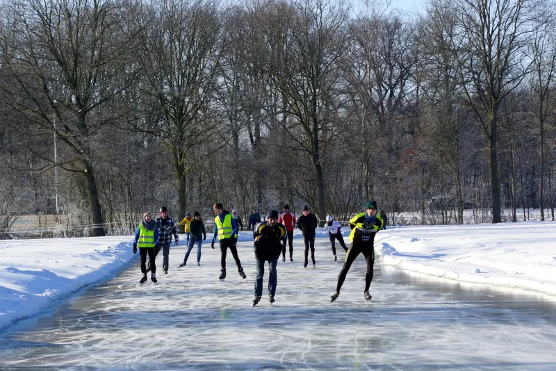 Een groep schaatsers in fluorescerende hesjes glijdt over de natuurijsbaan op Sportpark Zuid in Aalten, met sneeuwbermen en kale bomen erachter. – IJsvereniging de Hare