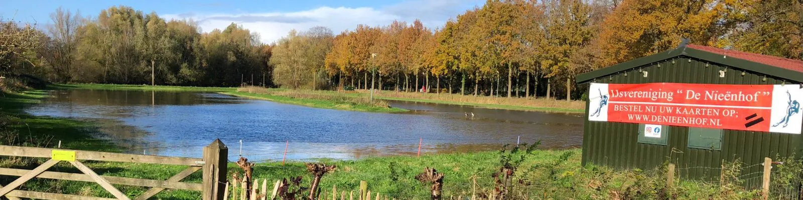 The green clubhouse hut of IJsvereniging De Nieënhof with its red club sign beside the flooded field, with autumn trees along the far edge. — IJsbaan De Nieënhof (IJsvereniging De Nieënhof, Bunnik)