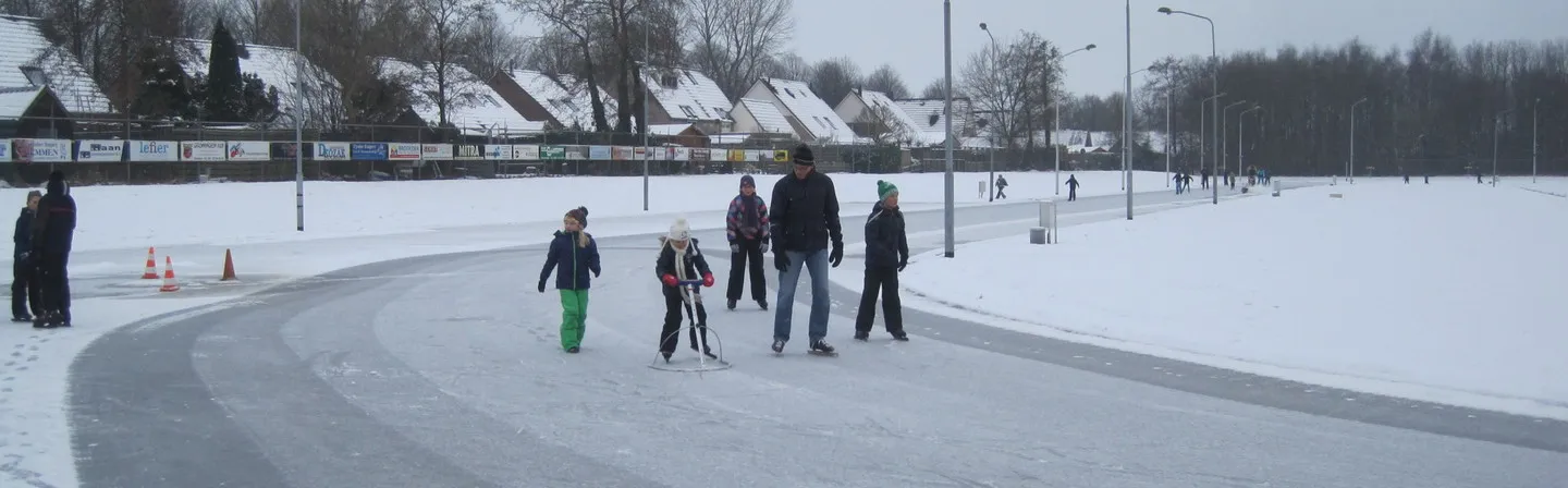 De natuurijsbaan in Alteveer in de winter, een geveegde grijze baan door de sneeuw met een paar schaatsers, lichtmasten en een rij huizen aan de overkant. – IJsbaan Alteveer (IJsvereniging 'De Toekomst')