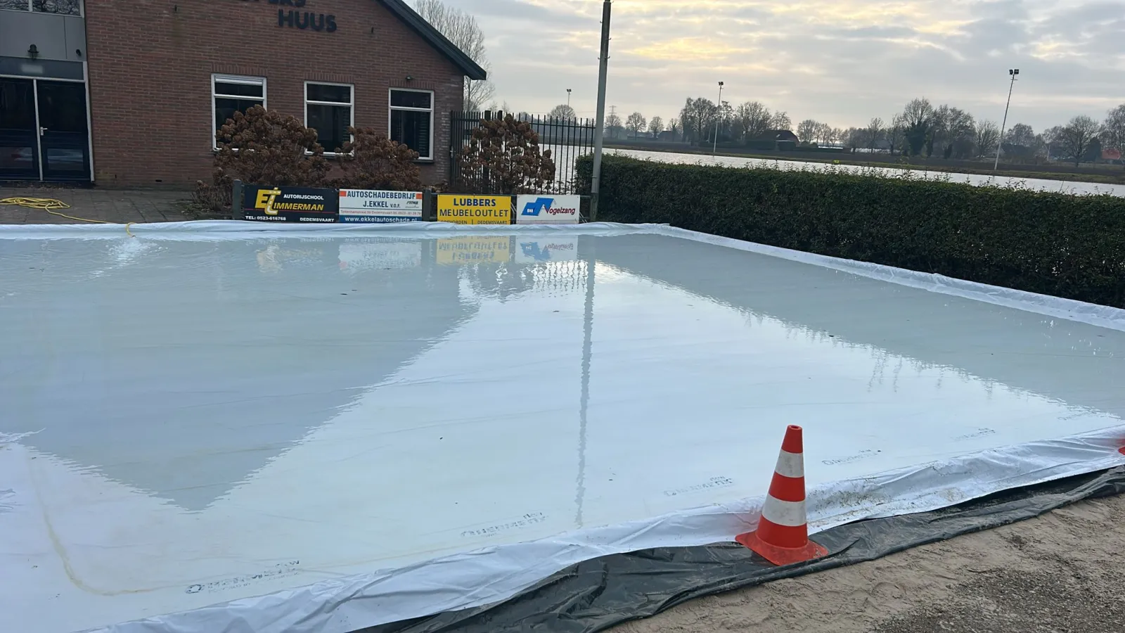 The small scrub rink for the youngest skaters laid out on a black liner in front of the brick Deurlopershuus clubhouse at Dedemsvaart, with orange cones and club sponsor boards along the edge. — IJsbaan Dedemsvaart (IJsvereniging Sluis 6-7)