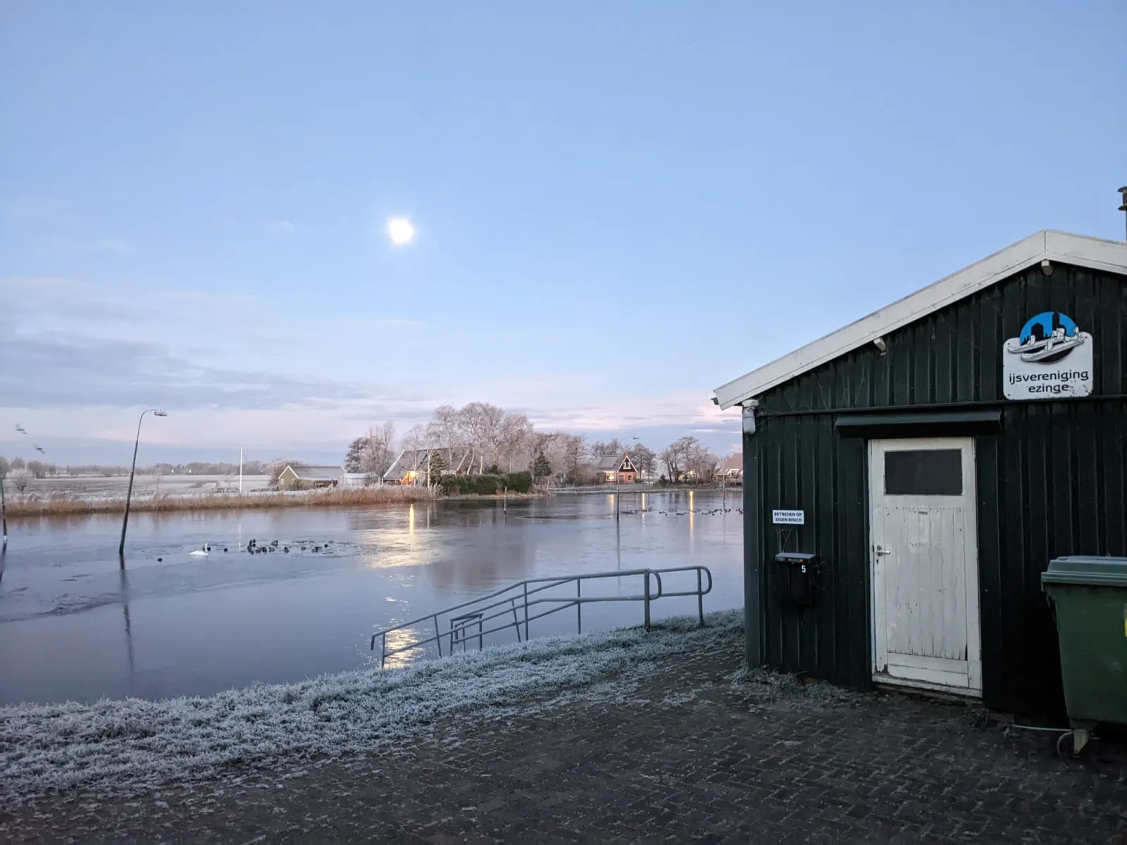 Het donkergroene clubhokje van IJsvereniging Ezinge met het clubbord op de gevel, naast de bevroren baan in de schemering met een hek en bomen langs het water erachter. – IJsbaan Ezinge (IJsvereniging Ezinge)
