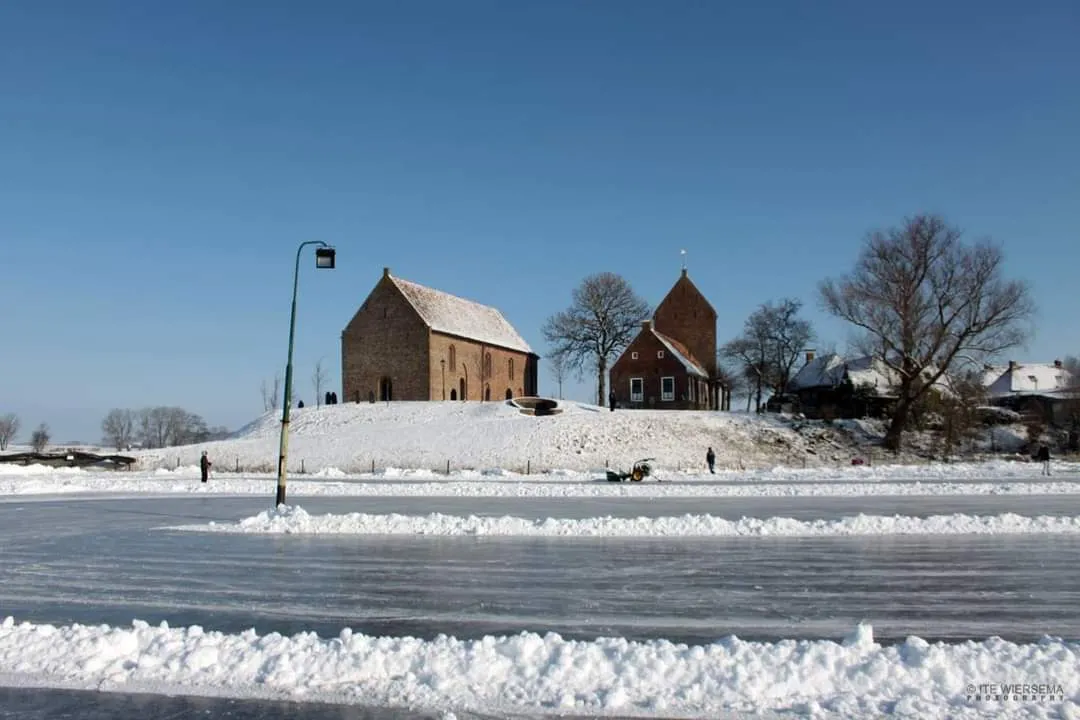De geveegde natuurijsbaan van Ezinge in de winter, met de oude kerk op de wierde aan de overkant van het ijs, een lantaarnpaal en sneeuw langs de rand. – IJsbaan Ezinge (IJsvereniging Ezinge)