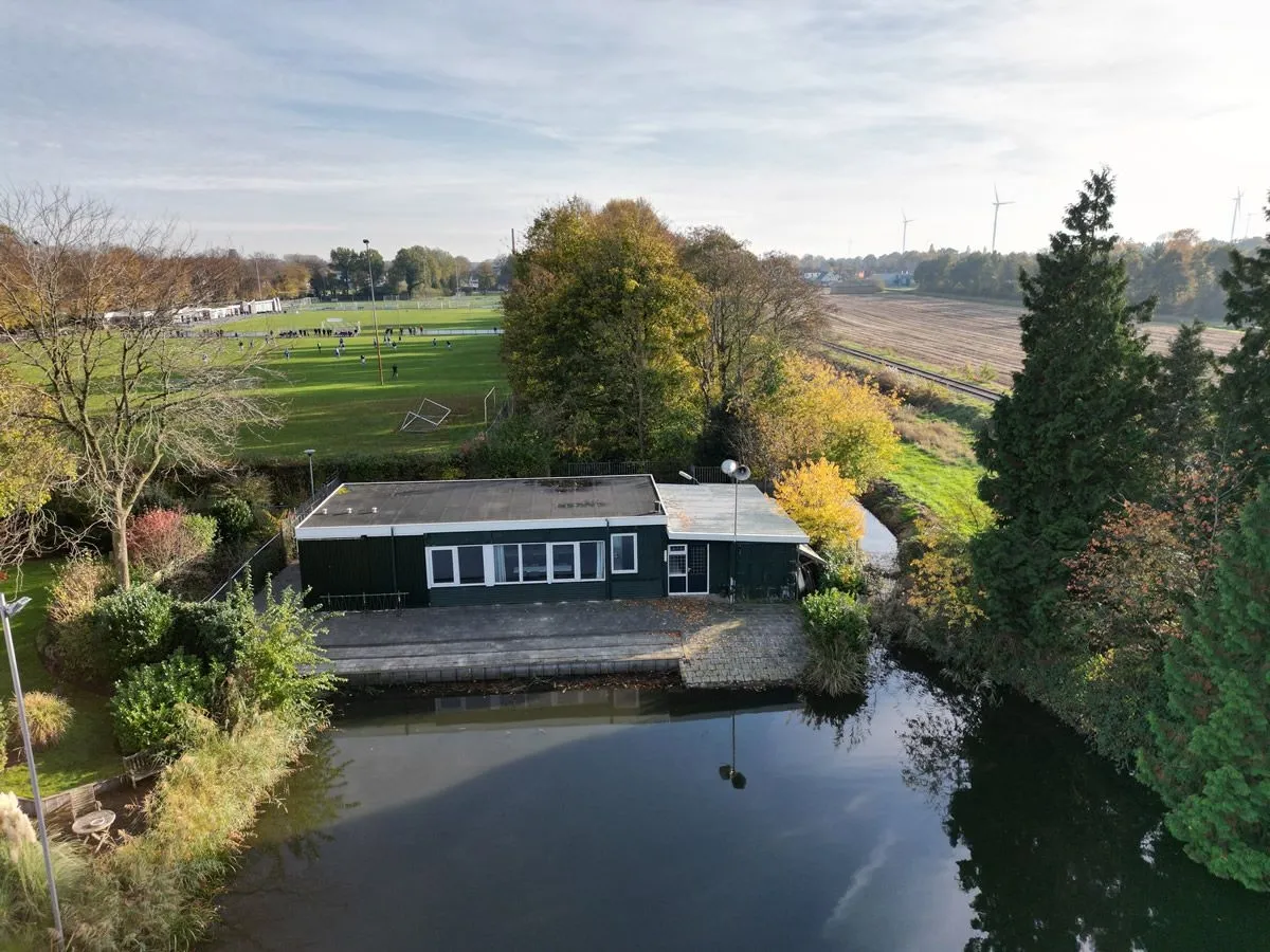 The dark-green low clubhouse of IJsvereniging Hamar beside the pond, seen from the air in autumn with trees and a sports field behind. — IJsbaan Hamar (IJsvereniging Hamar, Stadskanaal)