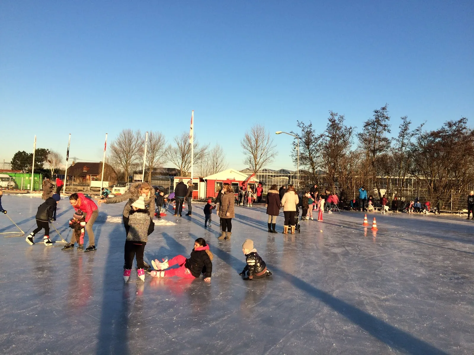 Schaatsers en kinderen op het open ijs van De Lier op een heldere winterdag, met vlaggenmasten, een witte tent en kale bomen achter de baan. – IJsvereniging Hard Gaat-ie (Piet Keijzerbaan)