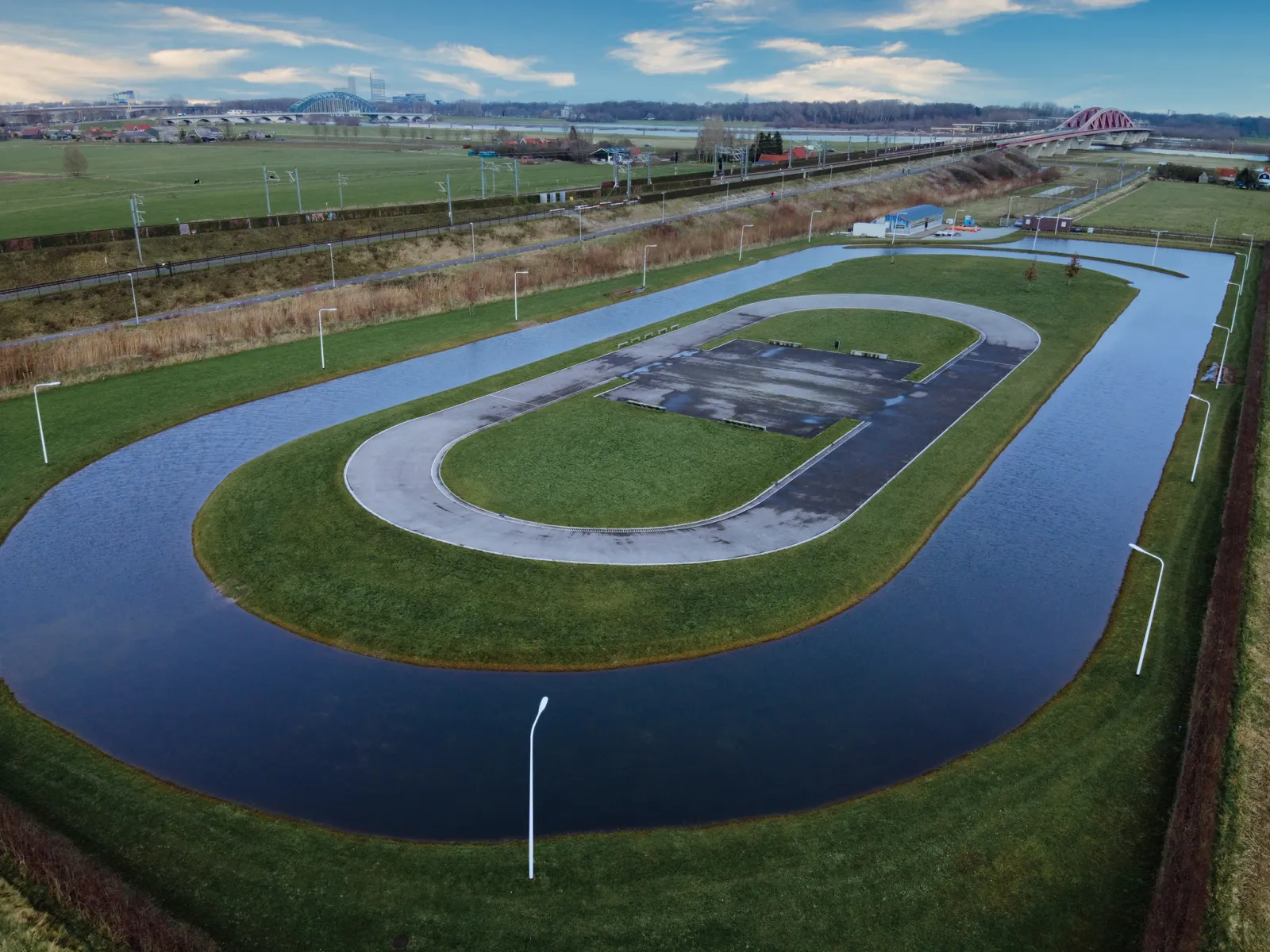Aerial view of the combined ice and skating track at Hattem on 25 January 2020, with the flooded land rink around the oval roller track, floodlights, the clubhouse building and the IJssel bridges behind. — IJsbaan Hattem (IJsvereniging Hattem)