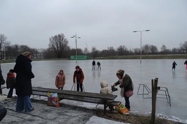 The natural ice of the IJsbaan Hoogkerk on a grey winter day, with a few skaters on the ice and families gathered by the bench at the edge. — IJsbaan Hoogkerk