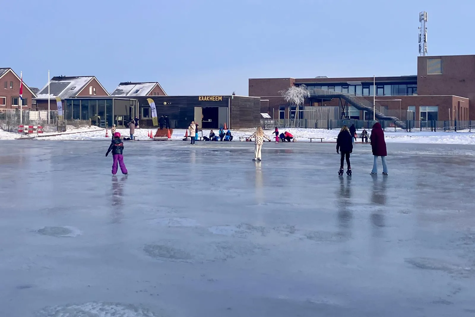 The Kaakheem club building and the Kindcentrum behind the frozen rink on a winter morning, with skaters on the blue-grey ice. — IJsbaan Kaakheem (IJsvereniging Kaakheem)