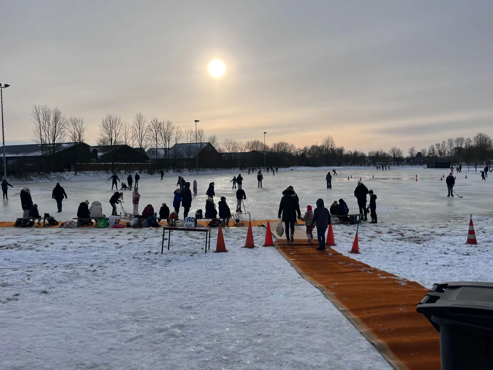A crowd of skaters and families on the natural ice of the Ten Boer rink at low sun, with a wooden walkway and cones along the edge and the town behind. — IJsbaan Kaakheem (IJsvereniging Kaakheem)