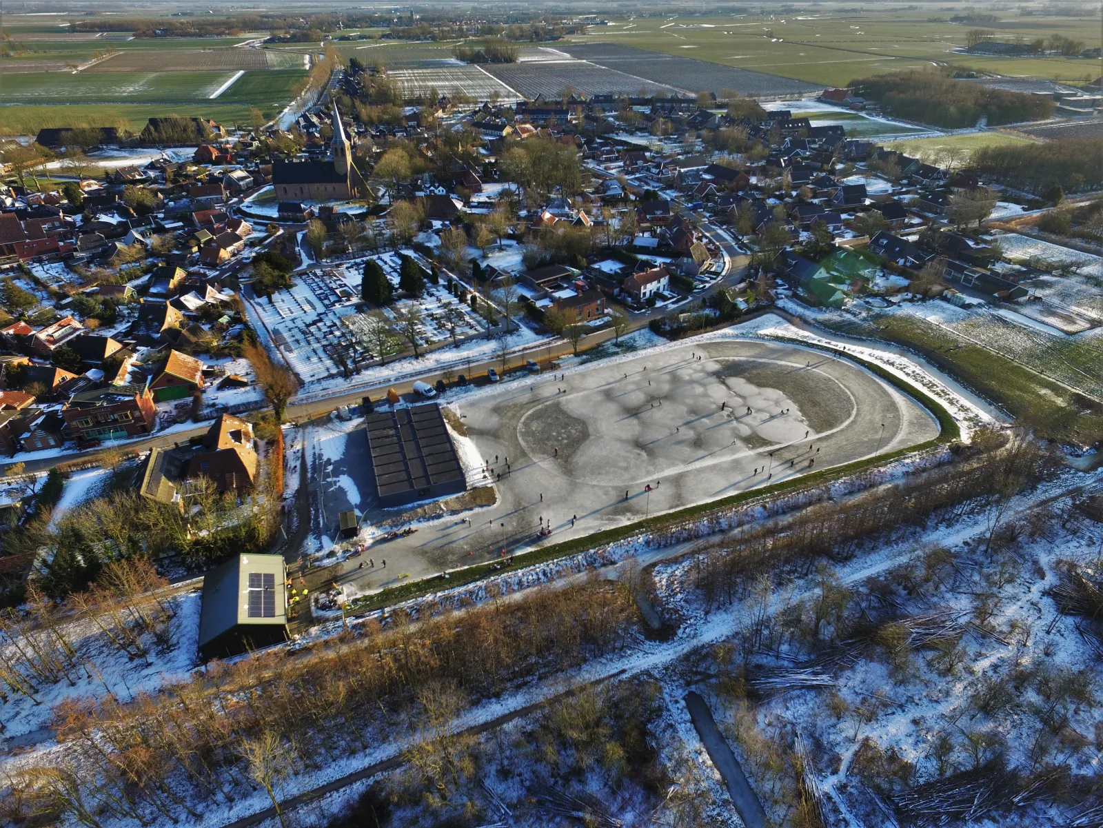 Aerial view of the Kantens village rink on a winter day, with skaters spread over the oval of ice beside the road, the church and snowy houses behind. — Thomas Wierema ijsbaan (Kantens)
