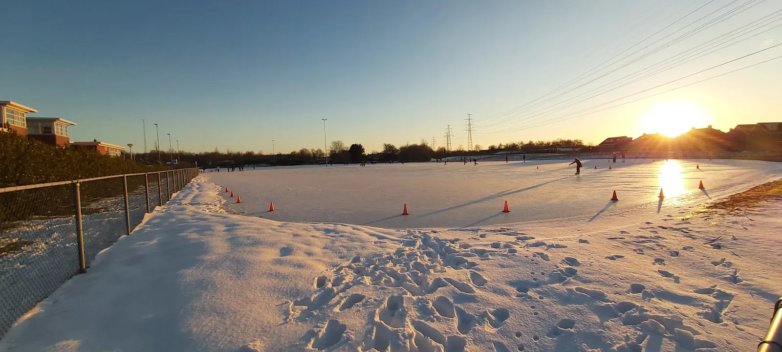 The snow-edged Kloosterveen natural-ice rink in low winter sun with orange cones marking the track and a few skaters far away. — IJsbaan Kloosterveen (IJsvereniging Kloosterveen)