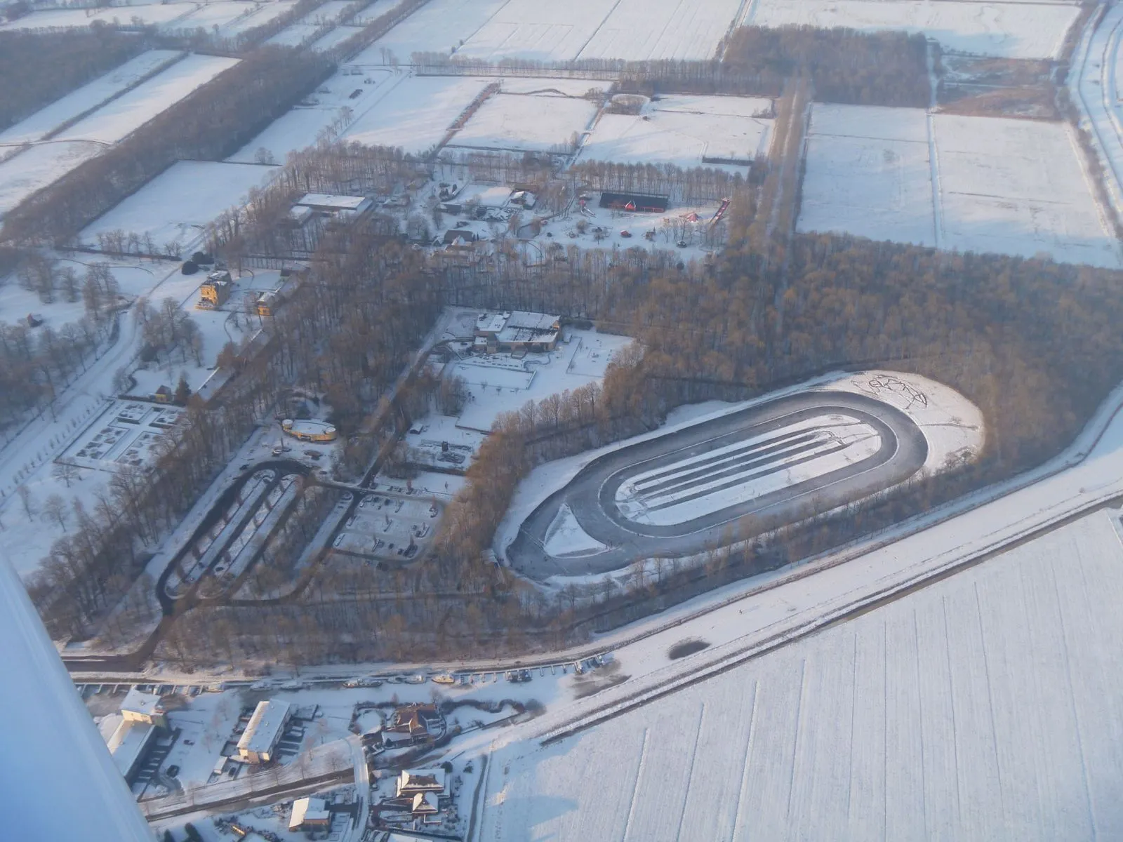 Een luchtfoto van de ovale natuurijsbaan van 400 m op landgoed Nienoord in de winter, omringd door bos en besneeuwde velden. – IJsbaan Nienoord (IJsvereniging Leek)