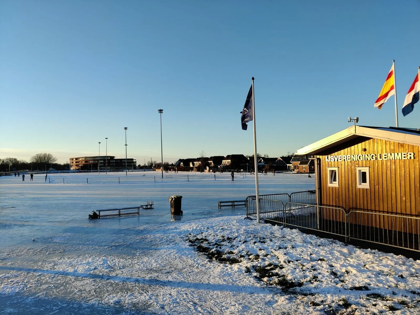 The timber clubhouse of IJsvereniging Lemmer with its name on the wall, flags on the mast and the smooth ice stretching away behind. — IJsvereniging Lemmer