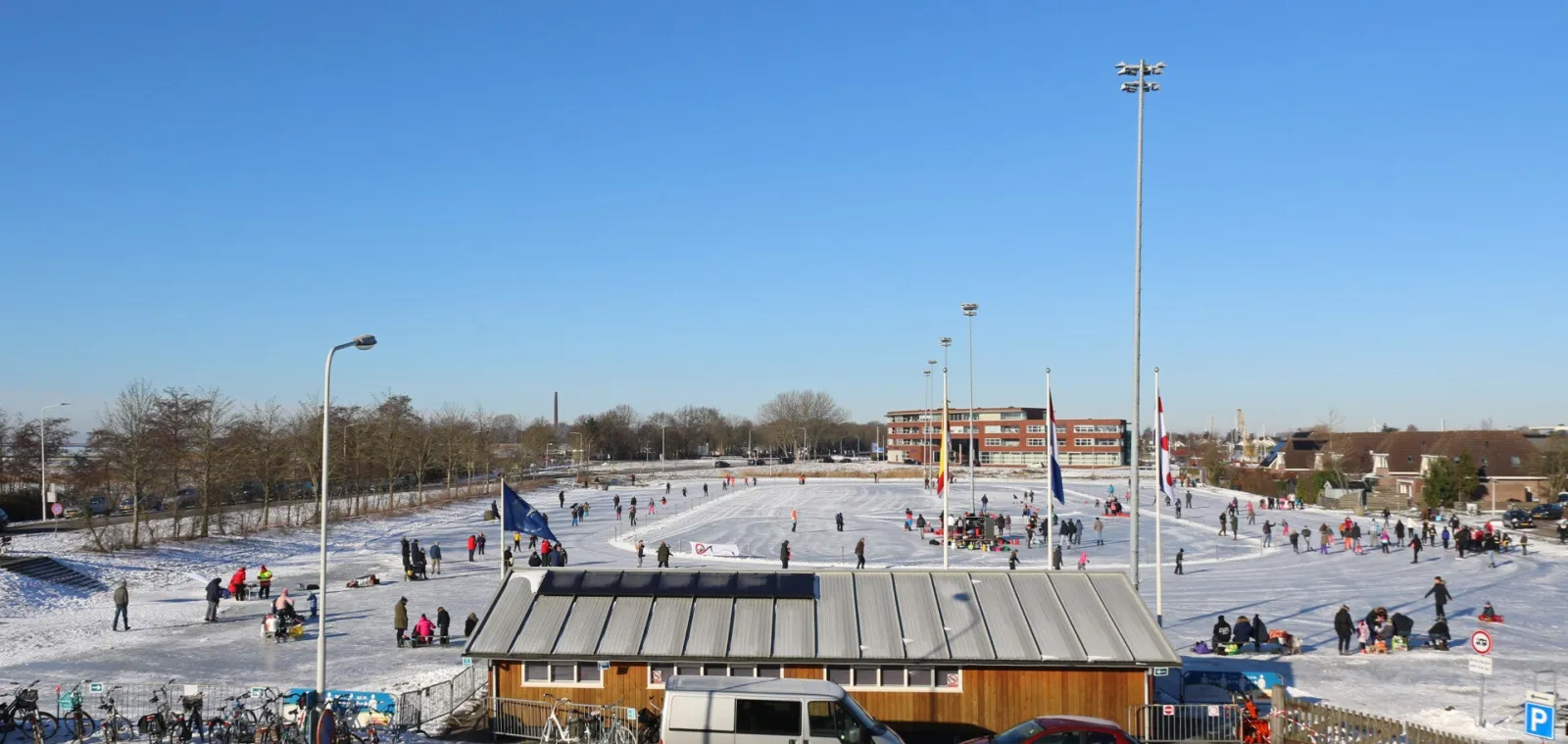 Skaters spread across the frozen Lemmer rink on a sunny winter day, with floodlight masts, flags and the timber clubhouse at the centre. — IJsvereniging Lemmer