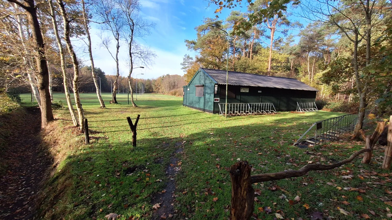 The dark green clubhouse hut on the meadow beside the rink in autumn, framed by birches and pines. — IJsbaan Molenveen (Norg)