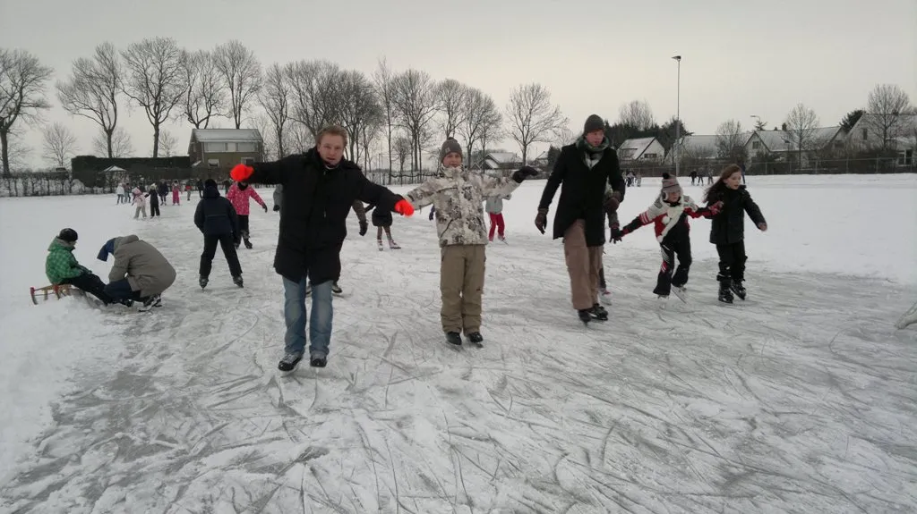 Skaters and families on the snow-dusted natural ice at Numansdorp in February 2012, with a sledge in the foreground and trees and houses along the far side. — IJsbaan Nooitgedagt (Numansdorp)