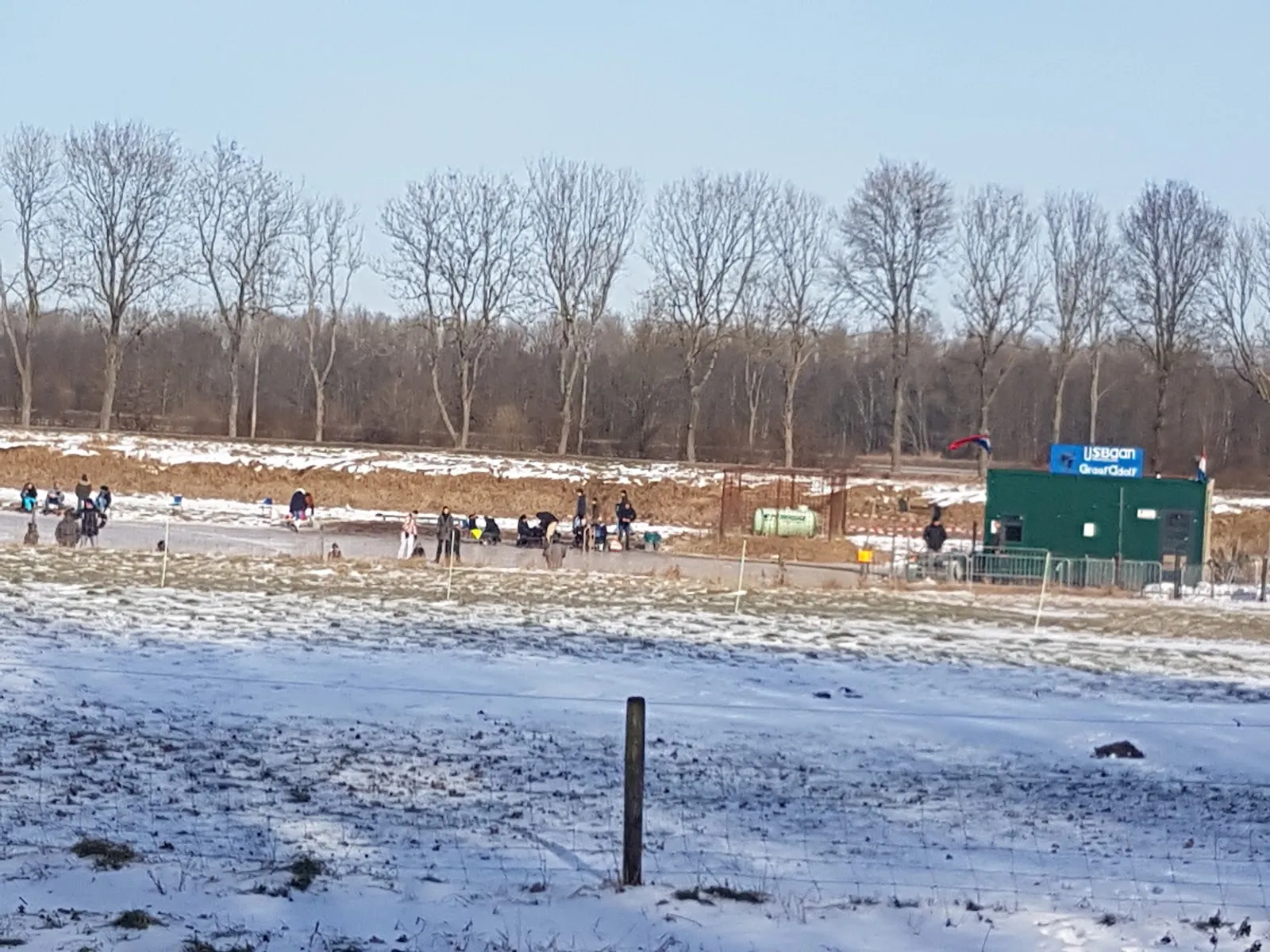 De met sneeuw bestoven baan van Scheemda met het groene kioskhuisje en een paar schaatsers aan de overkant onder kale bomen. – IJsvereniging Oldambt