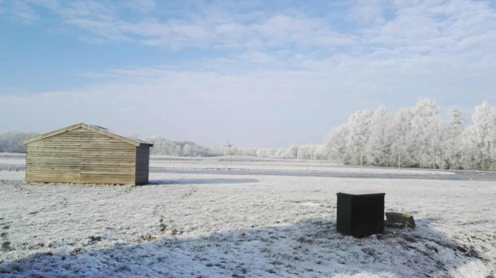 Een houten schuur op het berijpte baanterrein van Gasselternijveen onder een bleke winterlucht, met berijpte bomen aan de overkant van het veld. – IJsvereniging Oostermoer