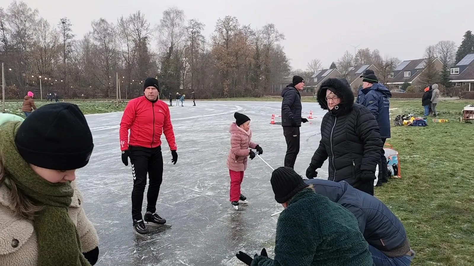 Schaatsers en gezinnen op de verlichte natuurijsbaan van Gasselternijveen op een winterse middag, met kale bomen en huizen achter de rand van het ijs. – IJsvereniging Oostermoer