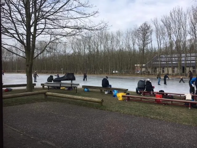 Uitzicht over het natuurijsveld van Pijnacker vanaf de oever, met bankjes op de voorgrond, schaatsers op het ijs en het clubgebouw de Tinteltent en kale bomen aan de overkant. – Natuurijsbaan Pijnacker (IJsvereniging Pijnacker, Tinteltent)