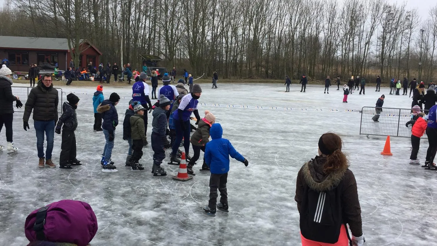 Een menigte kinderen en volwassenen schaatst op het grijze natuurijs van Pijnacker in februari 2018, met verkeerskegels, een clubgebouw en kale bomen langs de overkant. – Natuurijsbaan Pijnacker (IJsvereniging Pijnacker, Tinteltent)