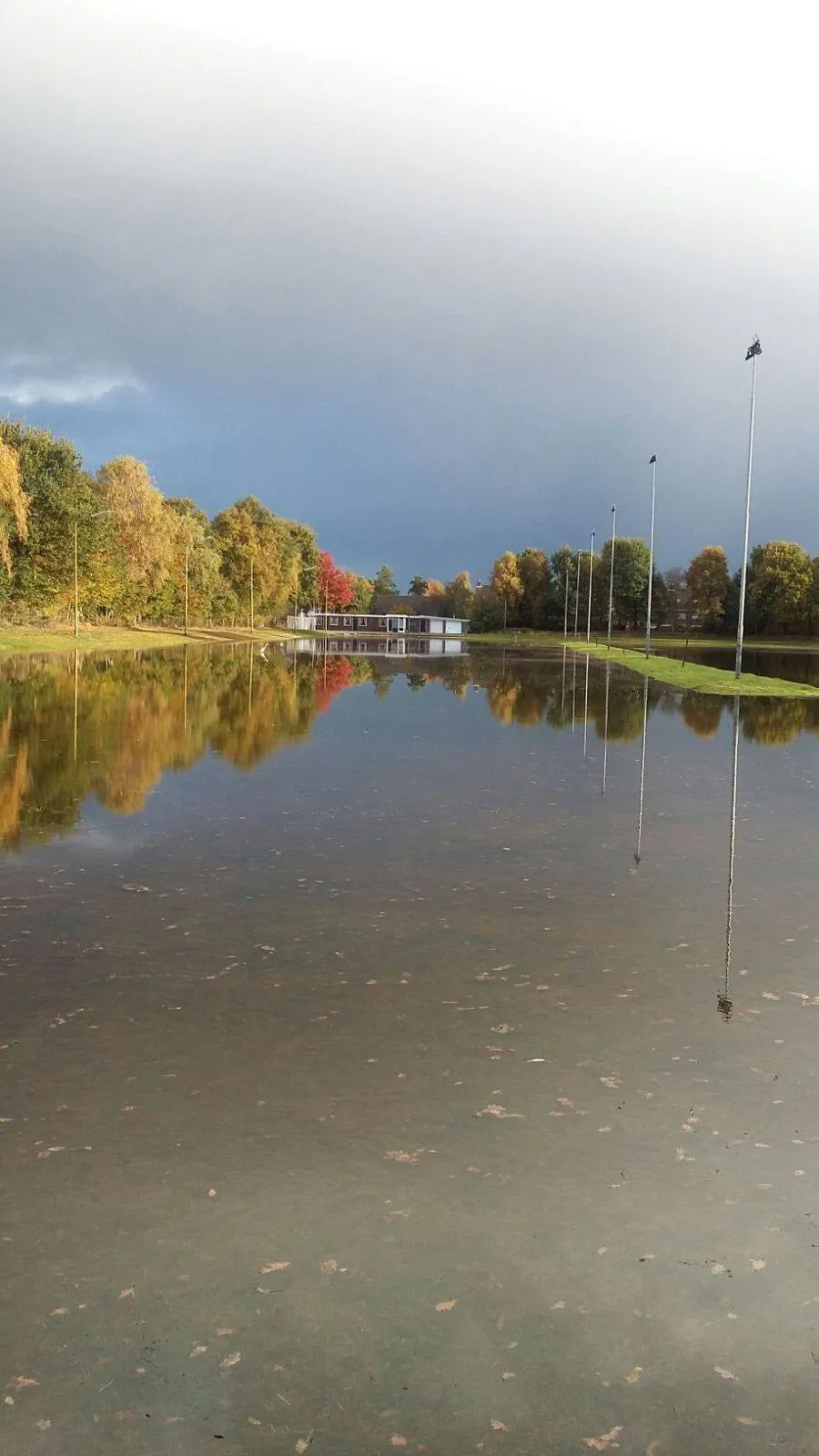 The flooded rink at Bovensmilde in autumn with the low white clubhouse across the water, flagpoles in a row and trees in autumn colour behind. — IJsbaan Bovensmilde (IJsvereniging Voorwaarts)