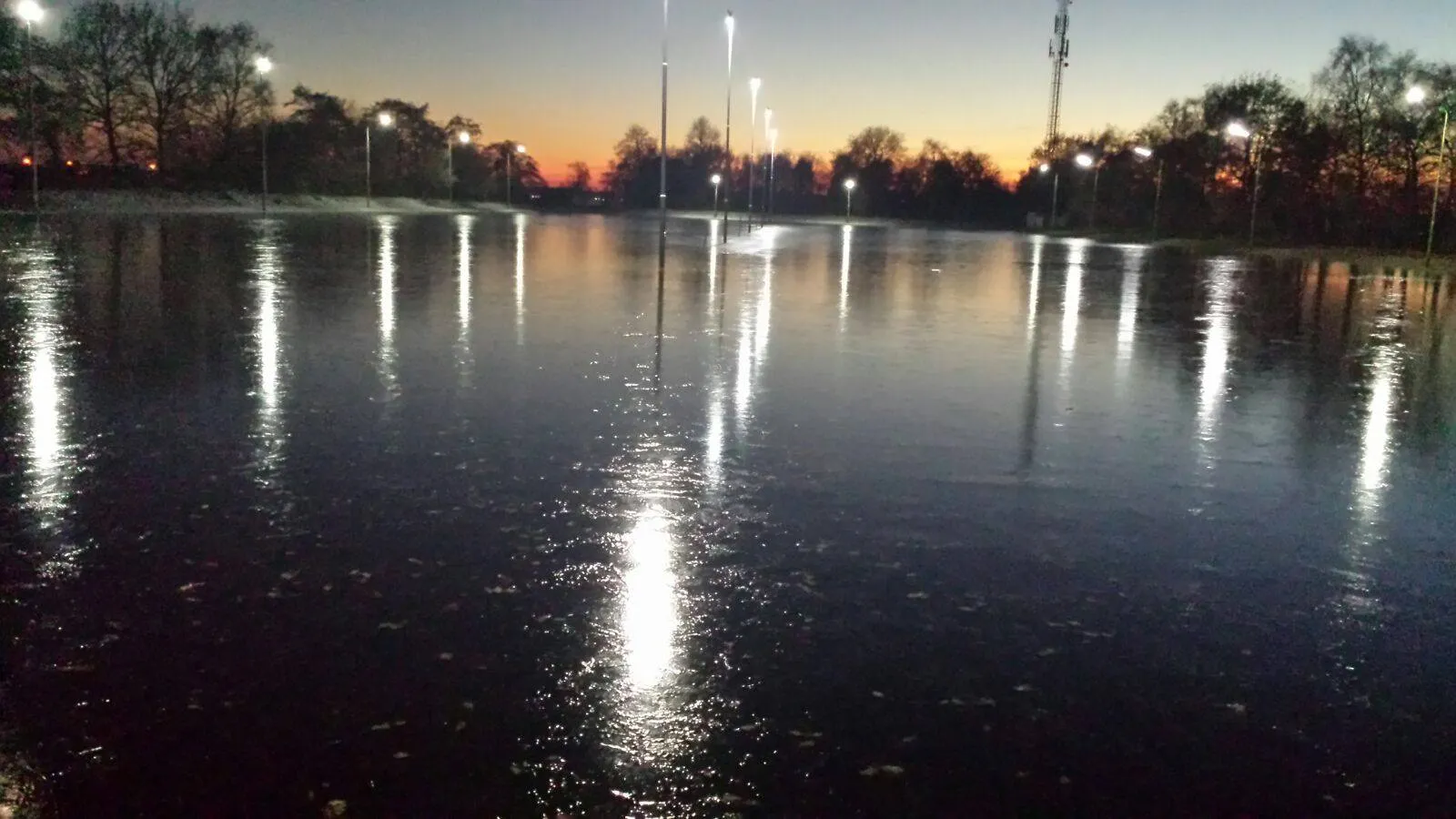 The floodlit natural-ice rink at Bovensmilde at dusk, a smooth dark ice surface with the lamps and bare trees reflected in it under an orange sky. — IJsbaan Bovensmilde (IJsvereniging Voorwaarts)