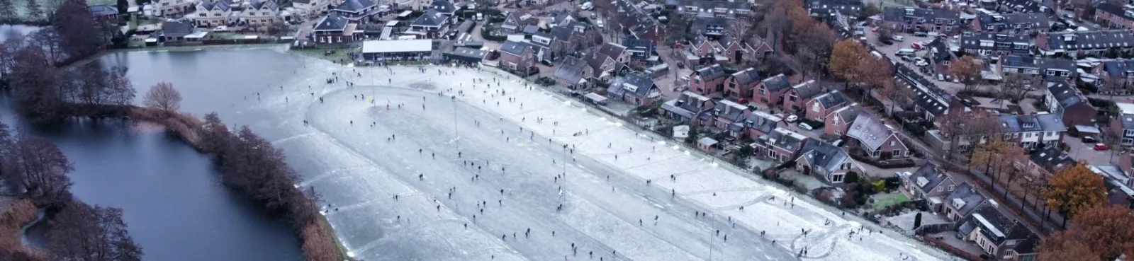 Luchtfoto van de WVF-baan in Zwolle, een lange strook ijs met schaatsers naast een waterloop en de rand van de woonhuizen. – IJsbaan WVF (IJsvereniging WVF)