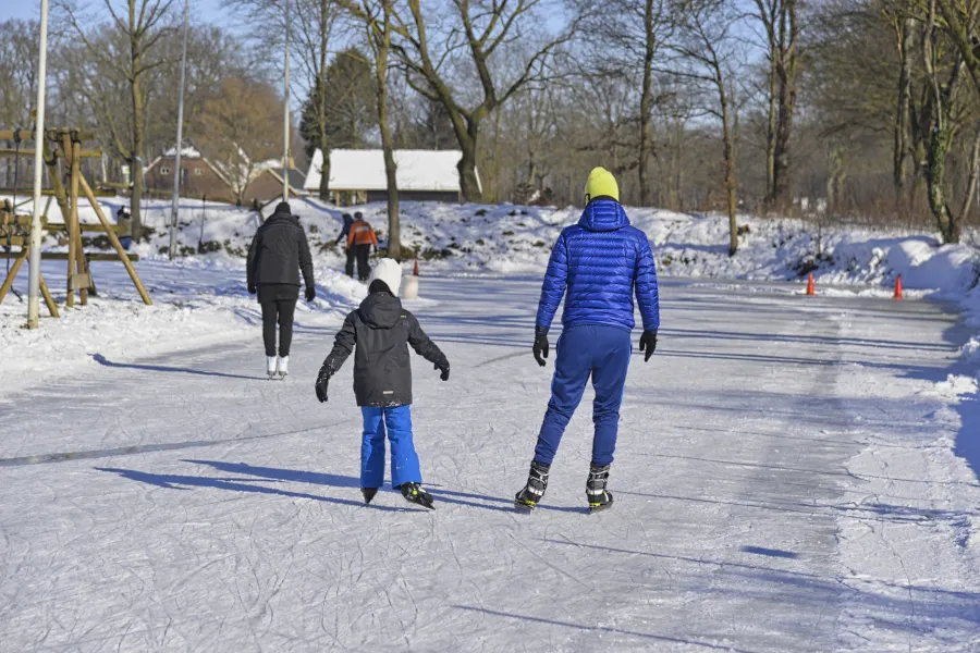 Schaatsers en kinderen op de besneeuwde natuurijsbaan van Zelhem, met kale bomen en het houten speeltoestel aan de rand van de baan. – IJsvereniging Zelhem