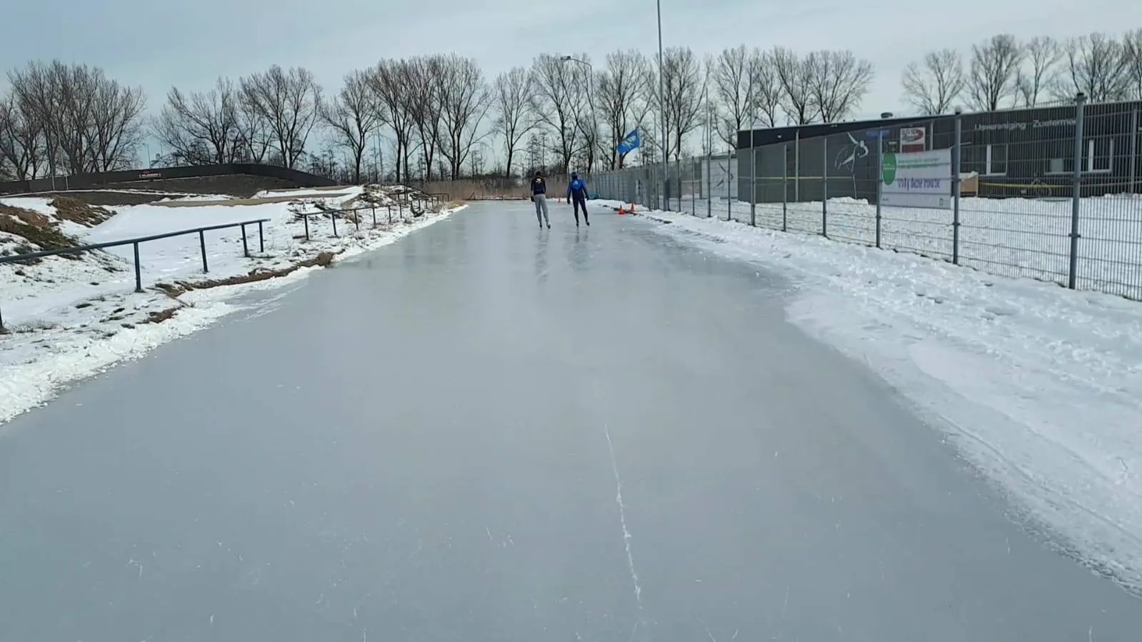 The flooded roller track at the Buytenpark in Zoetermeer frozen into a smooth grey ice lane in winter, with snow banked beside it, a mesh fence, bare trees and two skaters in the distance. — Natuurijsbaan YVZ (Buytenpark Zoetermeer)