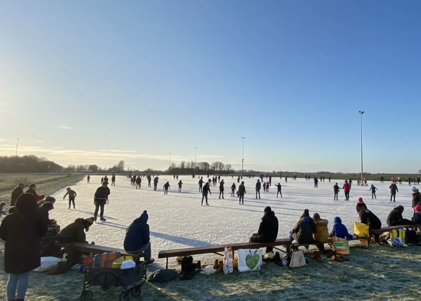 Schaatsers ver uit elkaar op het zonovergoten natuurijs van de Zuidwolder baan onder een diepblauwe lucht, met mensen die uitrusten op banken langs de voorste rand. – IJsbaan Zuidwolde (IJsvereniging Zuidwolde)