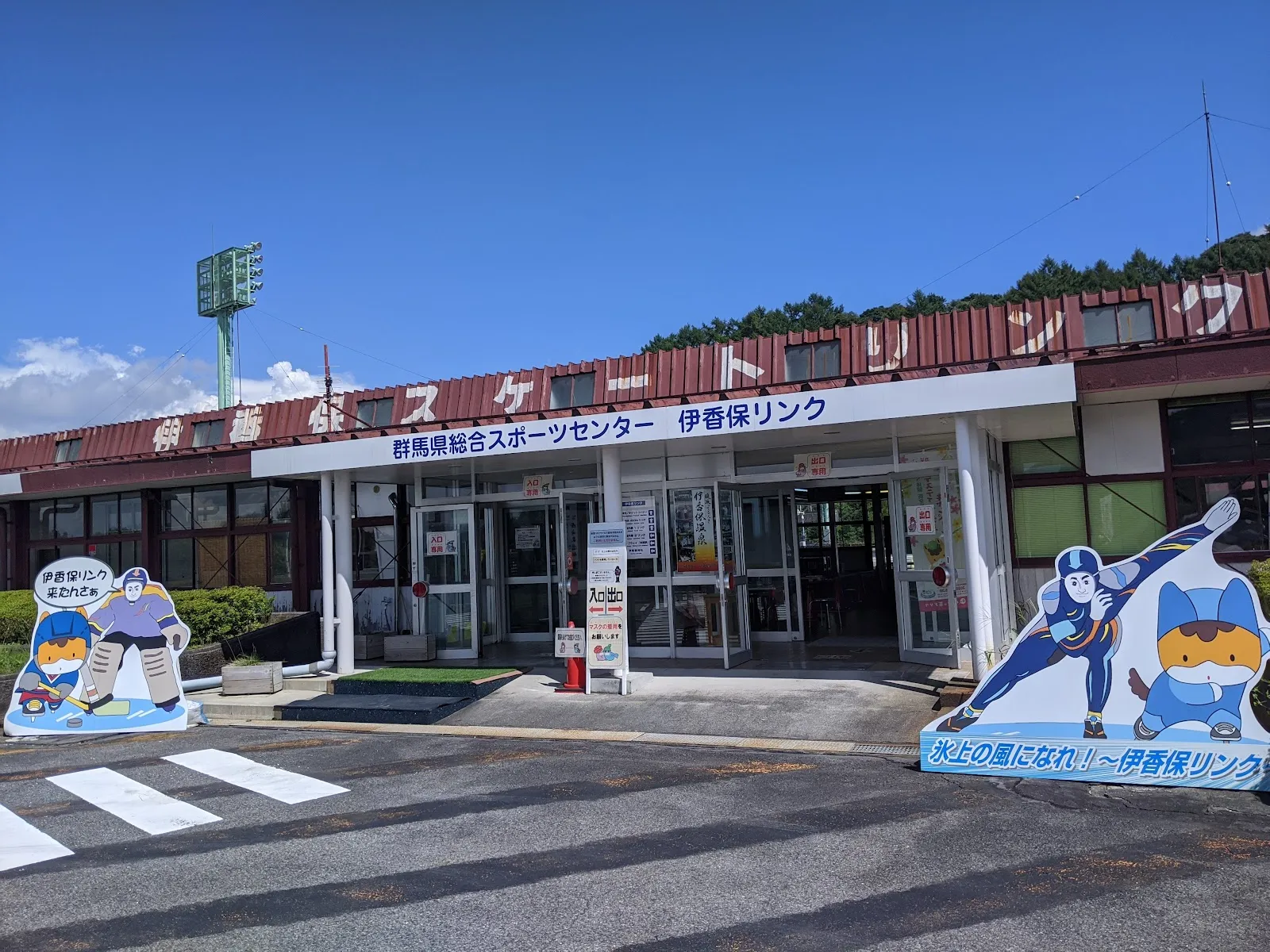 「群馬県総合スポーツセンター 伊香保リンク」の看板がある、赤い屋根の建物の下にある伊香保リンクの入口。両側に、スケーターとホッケー選手のマスコットのパネルがあります — Ikaho Rink (Gunma Sports Center)