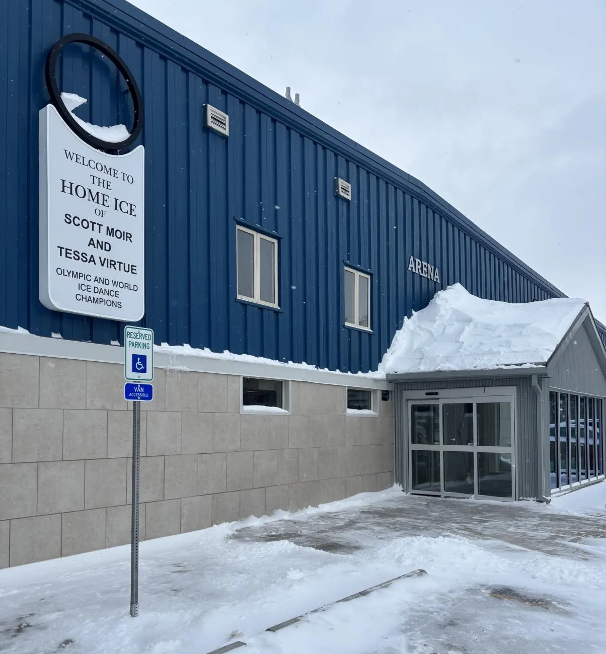 The blue-clad Ilderton Arena with its Home Ice of Scott Moir and Tessa Virtue welcome sign beside the glass entrance, snow on the walk. — Ilderton Arena