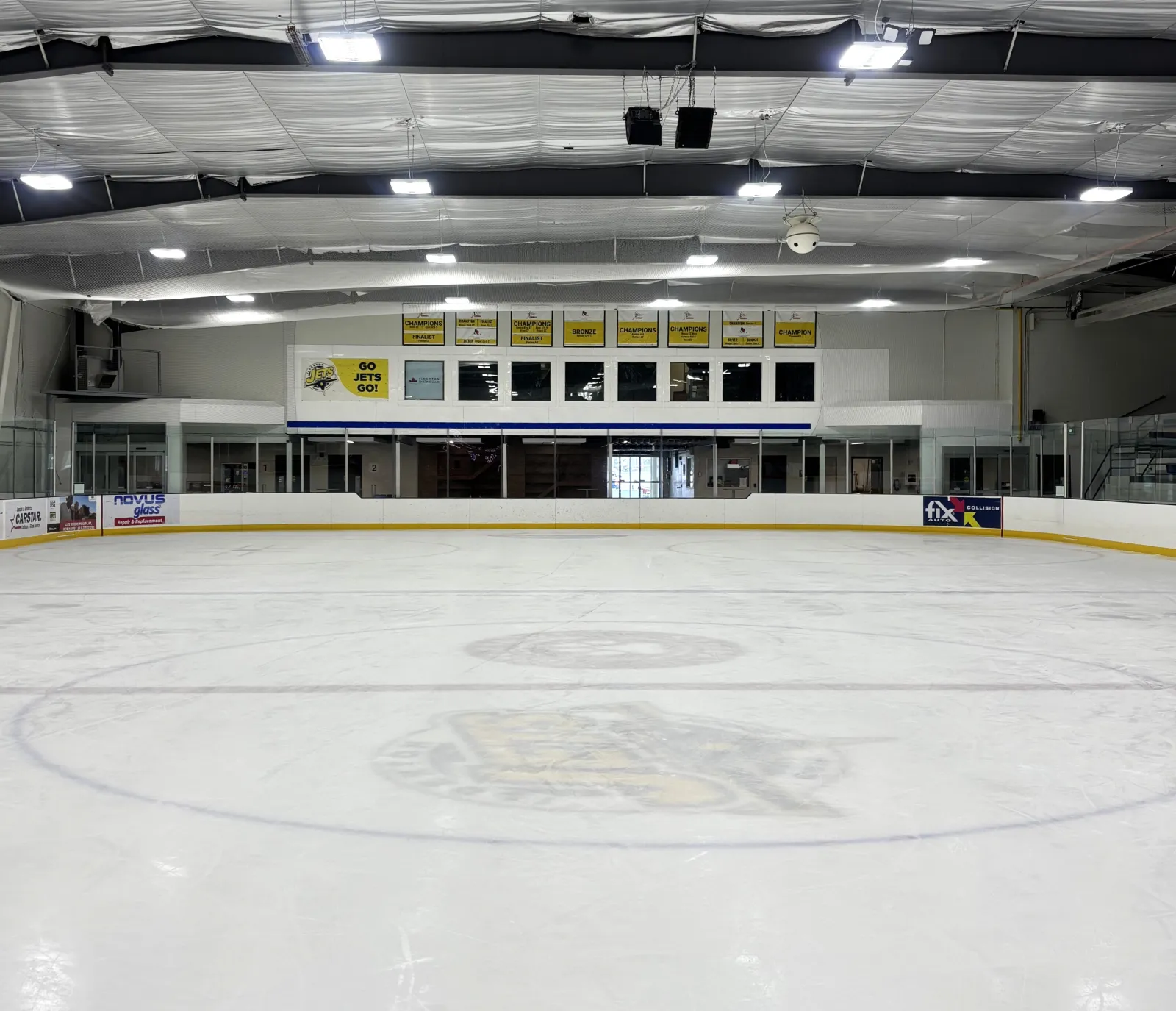 Ilderton Arena's ice under bright lights, with the centre-ice logo, the far-end seating and banners beyond the glass. — Ilderton Arena