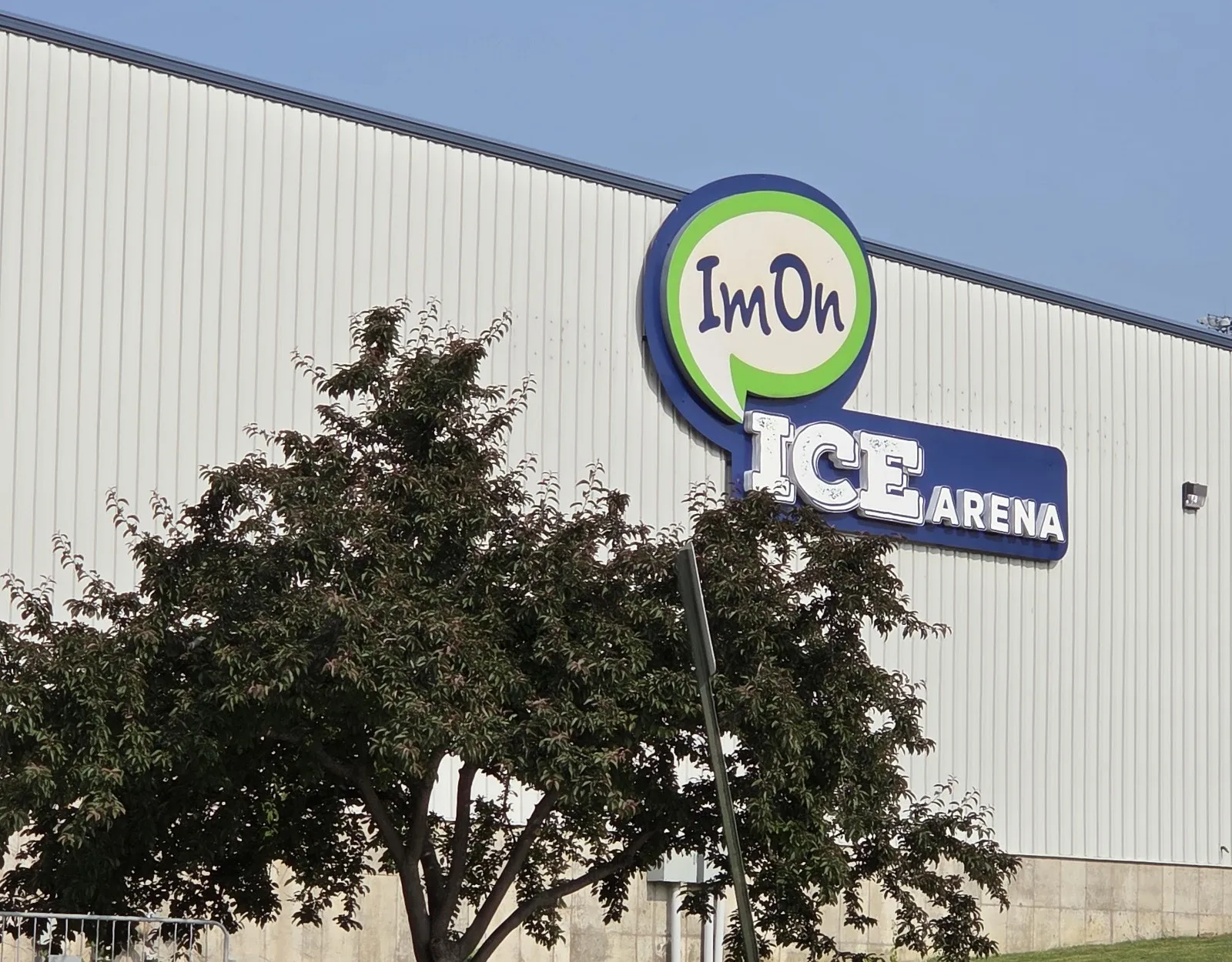 El letrero de ImOn Ice Arena en el largo muro metálico acanalado de la arena, con un árbol en flor y pasto al frente — ImOn Ice Arena (Cedar Rapids Ice Arena)