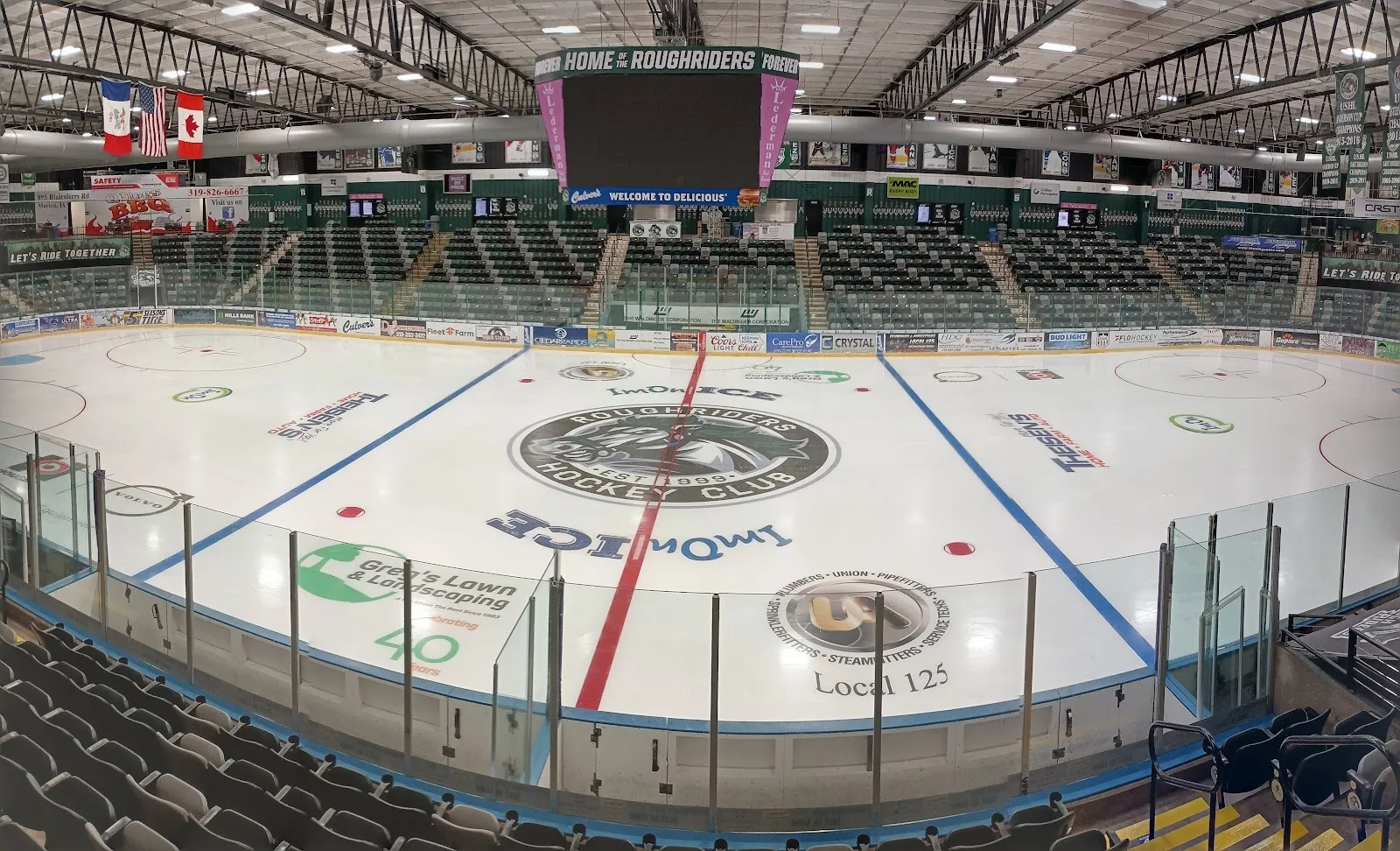 La arena vista desde las gradas, con el hielo vacío, un logotipo central y líneas azules y rojas bajo un marcador iluminado y un techo de acero. — ImOn Ice Arena (Cedar Rapids Ice Arena)