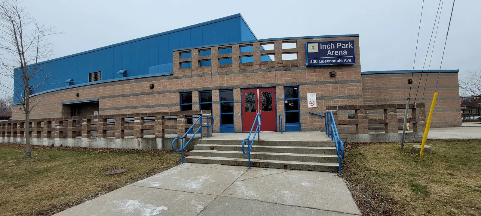 The front of Inch Park Arena in Hamilton, a brick building with a blue roof, red entrance doors and the arena sign at 400 Queensdale Avenue — Inch Park Arena