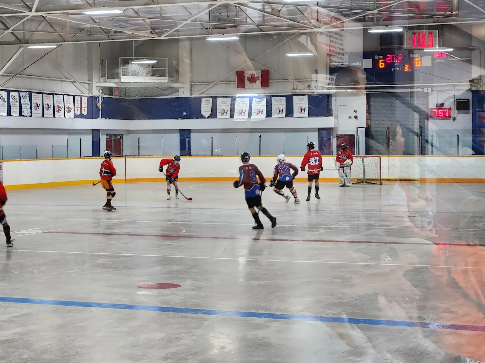 A practice on a bright sheet under a steel-trussed roof, players in red and navy spread across the far end with a blue line along the near boards. — Inch Park Arena